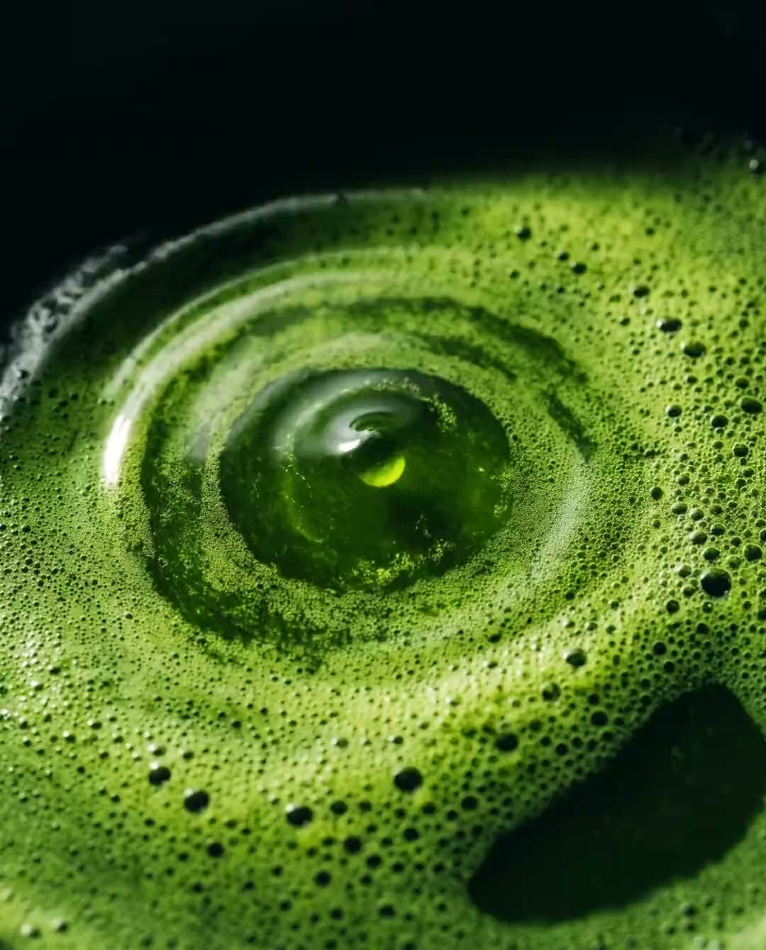 A slow ripple crossing the surface of whisked matcha, lit from one side against near-black.
